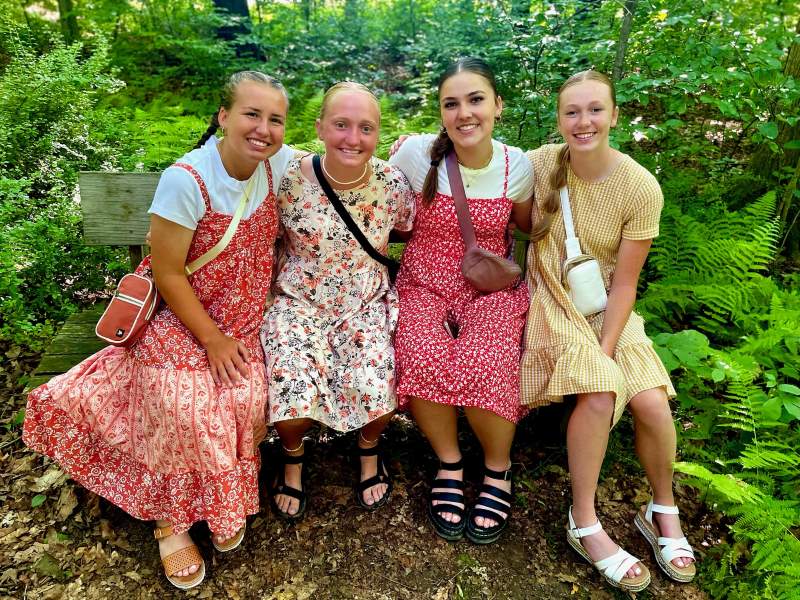 Four young women in Sunday dresses smile while sitting on a bench in a wooded area.